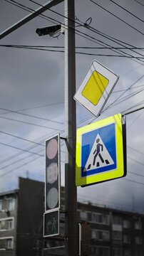 Urban intersection with pedestrian crosswalk sign, inactive traffic lights, priority warning symbol, surveillance camera, and overhead wires against a cloudy city skyline