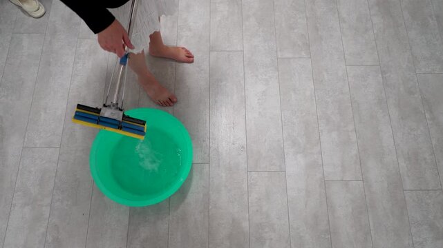 A person is using a damp mop to clean wooden floors, standing barefoot next to a bowl of water. The floor has a light, modern design, and the atmosphere is bright and cheerful.