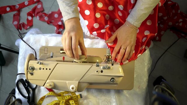 A person skillfully sews a vibrant red polka dot dress using a classic sewing machine. Measuring tape and fabric remnants are present, indicating ongoing creative work.