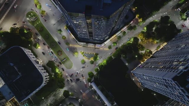 Aerial top view of a Hanoi crossroad surrounded by high-rise buildings and tree-lined streets, illuminated at night with moderate traffic movement and structured layout.