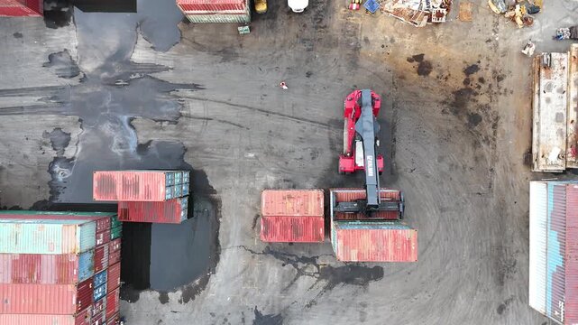High-angle aerial drone shot of a busy logistics terminal featuring a reach stacker crane moving industrial shipping containers in a large storage yard for global import export trade operations.