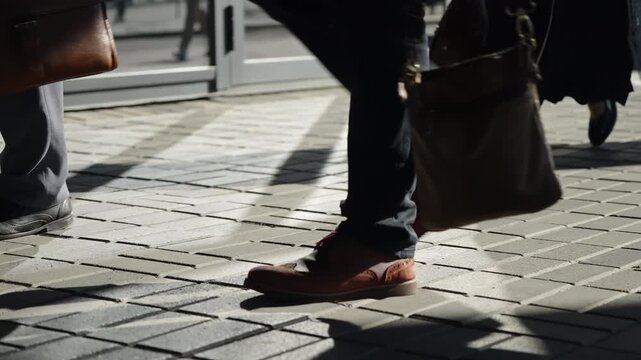 Urban commuters stride past a modern building entrance on a sunlit sidewalk, captured from ground level with focus on legs, shoes, briefcases, and dynamic shadows during a busy rush