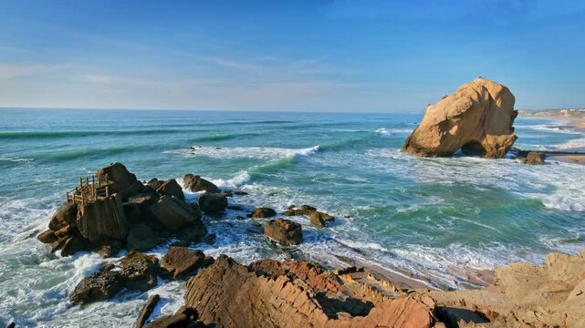 Penedo do Guincho stands at Praia da Santa Cruz, Portugal. Waves crash against rocks while visitors watch the beach and ocean at sunset. A large rock arch looms over the shore.