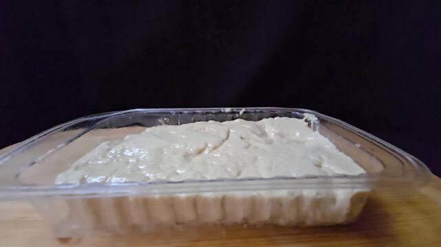 Sesame halva in a transparent plastic container. Close-up side view of traditional Mediterranean sweet in retail packaging on a wooden surface.