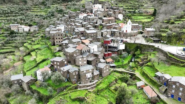 Aerial view of the village with stone houses, and a white church, nestled within vibrant green terraced hillsides, Piodao, Arganil, Portugal.