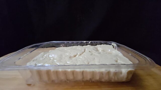 Sesame halva in a transparent plastic container. Close-up side view of traditional Mediterranean sweet in retail packaging on a wooden surface.
