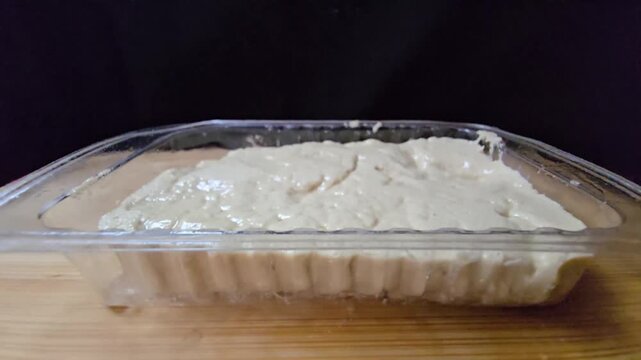 Sesame halva in a transparent plastic container. Close-up side view of traditional Mediterranean sweet in retail packaging on a wooden surface.