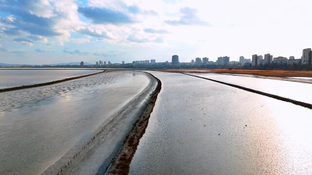 Aerial view of salt evaporation ponds near coastal city. Wide drone shot of salt pans and water canals with a city skyline under a cloudy sky.