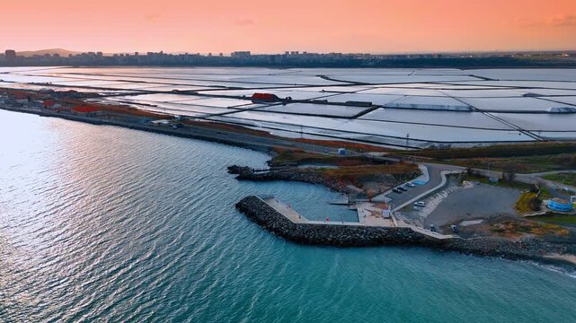 Aerial view of salt evaporation ponds near coastline at sunset. Salt pans separated by narrow paths under orange sky by the sea.