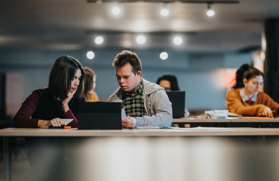 A tutor works with a young adult student with Down syndrome using a tablet while reviewing notes. The pair study together in a quiet, modern classroom setting with other students nearby.