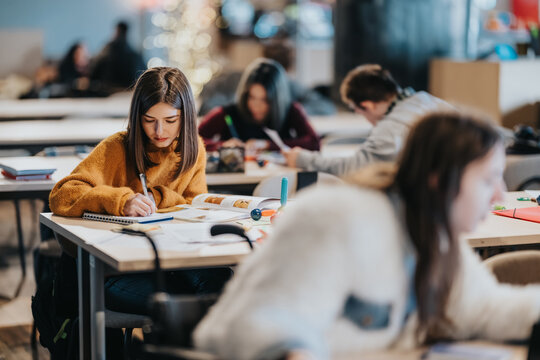 A young student studies intently at a desk surrounded by other students in a shared classroom. The scene shows a quiet group study environment with notebooks, pens and textbooks on tables.