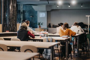 Obraz premium Adults study and collaborate at shared tables in a modern classroom or coworking workshop. A diverse group works with laptops, notebooks and art supplies while one person uses a wheelchair.