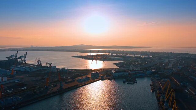 High angle view of maritime port under bright sun. Wide shot of harbor infrastructure, docks, and water reflection under a high sun. Clear sky and industrial zone.