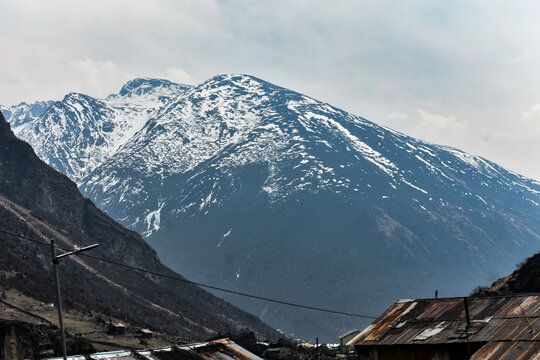 Rugged Himalayan valley with rocky terrain, scattered buildings, and distant snow peaks under dramatic cloudy sky in North Sikkim.