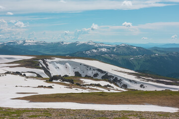 Scenic alpine landscape with sunlit snowy field on stony hill with view to big mountain range with forest and snow far away under cloudy sky. Stone outcrops among snows in sunlight in high mountains. © Daniil