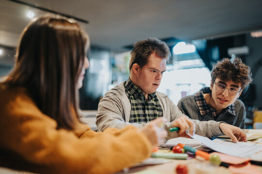 A group of young students studying together at a table, collaborating on papers and supplies. The scene shows focused, inclusive learning and peer support during a study session.