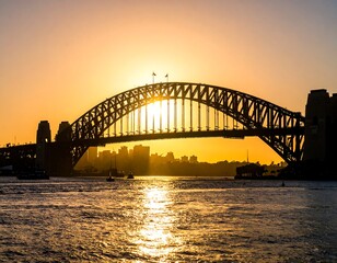 Fototapeta premium Silhouette of a large bridge spanning across water with a golden sunset in the background, city skyline visible