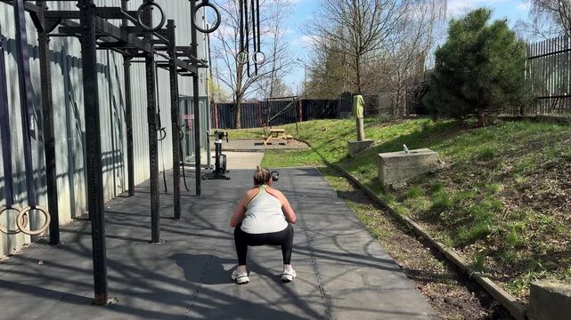 Woman performs an overhead medicine ball exercise at an outdoor functional fitness gym. Rear view sports scene with strength training, conditioning and active lifestyle in bright sunlight.
