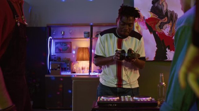 Young adult African American man using DJ controller playing music at home party while friends dancing together in living room