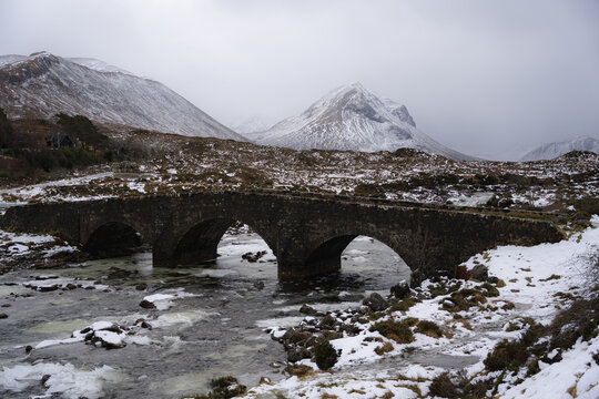 View of a Sligachan Old Bridge arches gracefully over a partially frozen river, with snow-dusted mountains rising in the background, Isle of Skye, Scotland.