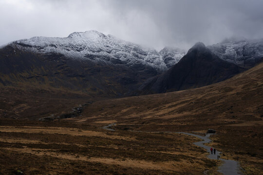 View of rugged Cuillin mountains capped with snow loom over a winding path where tiny figures stroll through the golden valley, Fairy Pools, Isle of Skye, Scotland.
