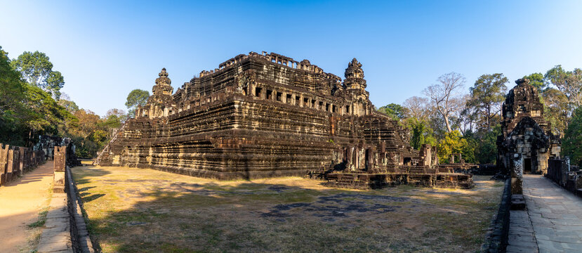 View of the Baphuon Temple imposing stone structure cuts a striking figure against the clear sky, framed by lush trees, Siem Reap, Siem Reap Province, Cambodia.