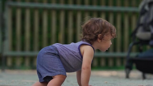 Toddler bending forward while sitting on playground ground studying sand closely, quiet childhood moment of curiosity observation and early discovery outdoors