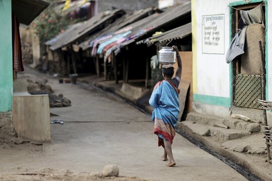 Odisha, India - 26 March 2026: View of a woman carrying water on her head along a dusty street lined with weathered homes, a scene of daily life.