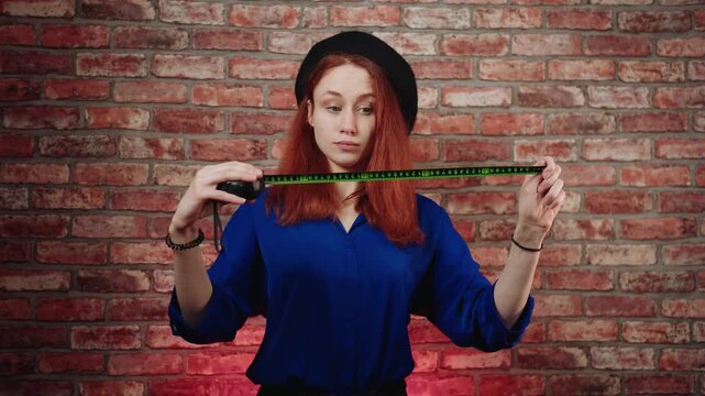 Front view of a caucasian young playful redhead woman in a blue blouse and black beret, opens the gauge tape and is surprised by the long length of the measuring tape against brick wall. Size matters