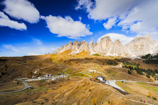 View of rugged, sunlit peaks pierce a vibrant blue sky dotted with fluffy clouds, casting shadows on the golden, rolling hillsides below, Gardena Pass, Trentino-South Tyrol, Italy.