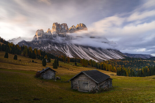 View of rustic wooden huts nestled in a valley beneath towering, jagged Odle-Geisler peaks partially veiled by soft, ethereal clouds, St. Magdalena, Trentino-South Tyrol, Italy.