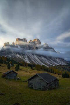 View of rugged Dolomites Odle peaks shrouded in misty veils, ancient wooden huts dotting the emerald slopes, St. Magdalena, Trentino-South Tyrol, Italy.