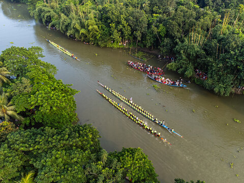 Aerial view of boats racing down a winding river, flanked by lush green trees, showcasing a vibrant display of tradition and nature, Gopalganj, Bangladesh.
