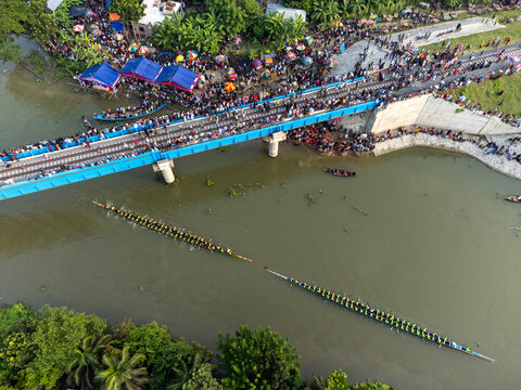 Aerial view of vibrant boat races cutting through the murky river under a crowded bridge, where spectators gather, Gopalganj, Dhaka Division, Bangladesh.