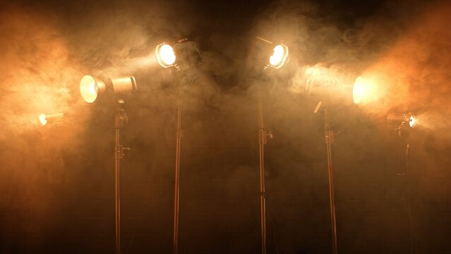 Multiple professional stage spotlights on stands shine in a dark smoke studio with visible beams. Concept of lighting equipment, theater production, concert setup, event technology and entertainment.