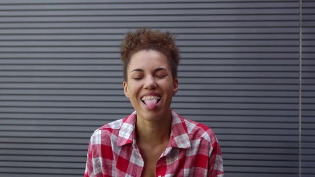 funny African-American woman sticks out her tongue and makes faces against urban wall, the girl teases and taunts the camera outdoors