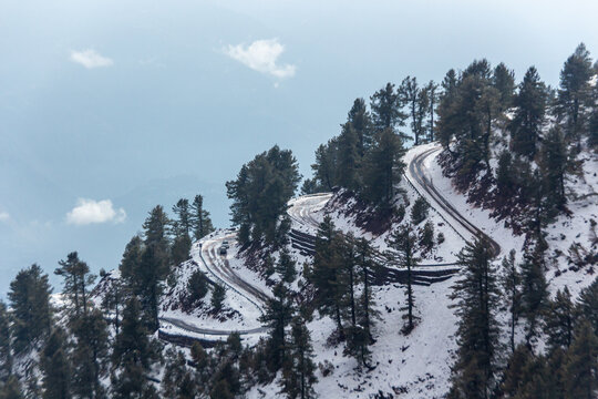 View of a winding road, cutting through a snow-dusted mountain embraced by towering pine trees under a soft sky, Pir Chanasi, Azad Kashmir, Pakistan.