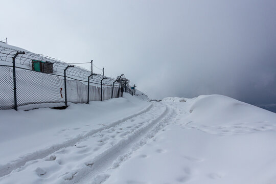 View of snow-covered ground with tire tracks leading towards a fence under a cloudy sky, Pir Chanasi, Azad Kashmir, Pakistan.