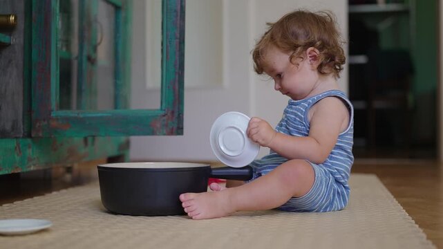 Toddler sitting on kitchen floor playing with cooking pot and small cup beside open cabinet exploring household objects during curious childhood moment at home