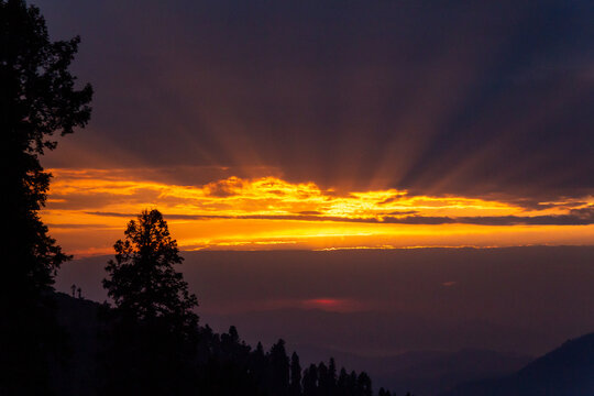 View of the sun's radiant rays pierce through dark, shadowy clouds over a silhouette of trees on a hillside, creating a dramatic scene, Pir Chanasi, Azad Kashmir, Pakistan.