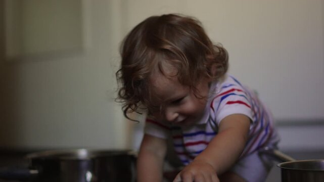 Toddler sitting on kitchen floor opening metal pot and lifting lid while exploring household objects with curiosity during everyday domestic play at home