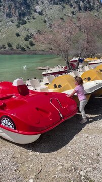 A curious little girl explores Lake Kournas in Crete, walking among colorful car-shaped catamarans. The scene features white geese on the turquoise water and mountains. High quality 4k footage
