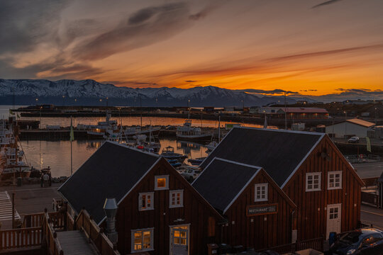 View of the harbor reflecting the fiery sunset sky, contrasted against the dark wooden buildings and distant snow-capped mountains, Husavik, Nordurthing, Iceland.