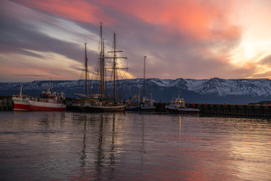 View of serene harbor reflecting a vibrant sunset sky with boats gently resting against the backdrop of snow-capped mountains, Husavik, Nordurthing, Iceland.