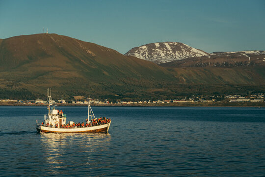 View of a boat brimming with passengers glides across the tranquil water, set against a backdrop of majestic mountains and a serene town, Husavik, Iceland.