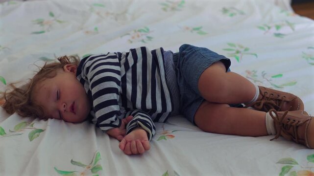 Toddler sleeping on bed in curled position with relaxed hand and small shoes, peaceful childhood rest moment inside quiet domestic family home bedroom
