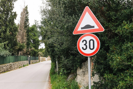 Road sign warning of speed bump with thirty limit on quiet street surrounded by greenery. Traffic safety sign placed near roadside. Caution and reduced speed zone in calm residential area with trees.