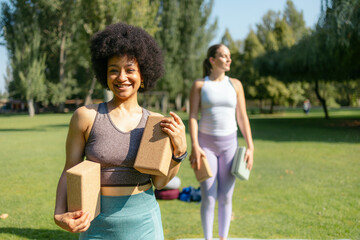 Two diverse women standing in a park, holding yoga blocks and smiling toward the camera