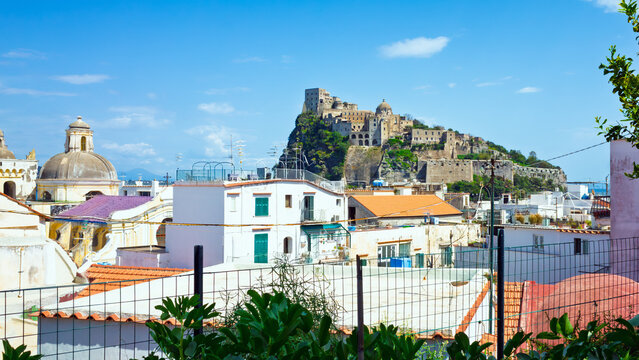 Aragonese Castle on rocky island overlooks coastal town rooftops on Ischia. Bright daylight on Ischia with blue sky and calm sea creates clear Mediterranean atmosphere