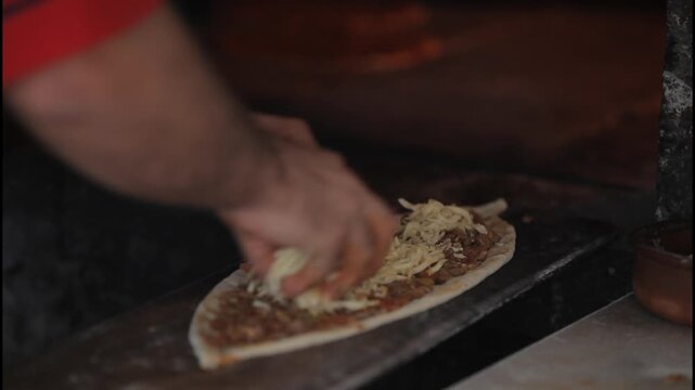 Adding shredded cheese to traditional Turkish pide and sliding it into a stone oven; static shot.